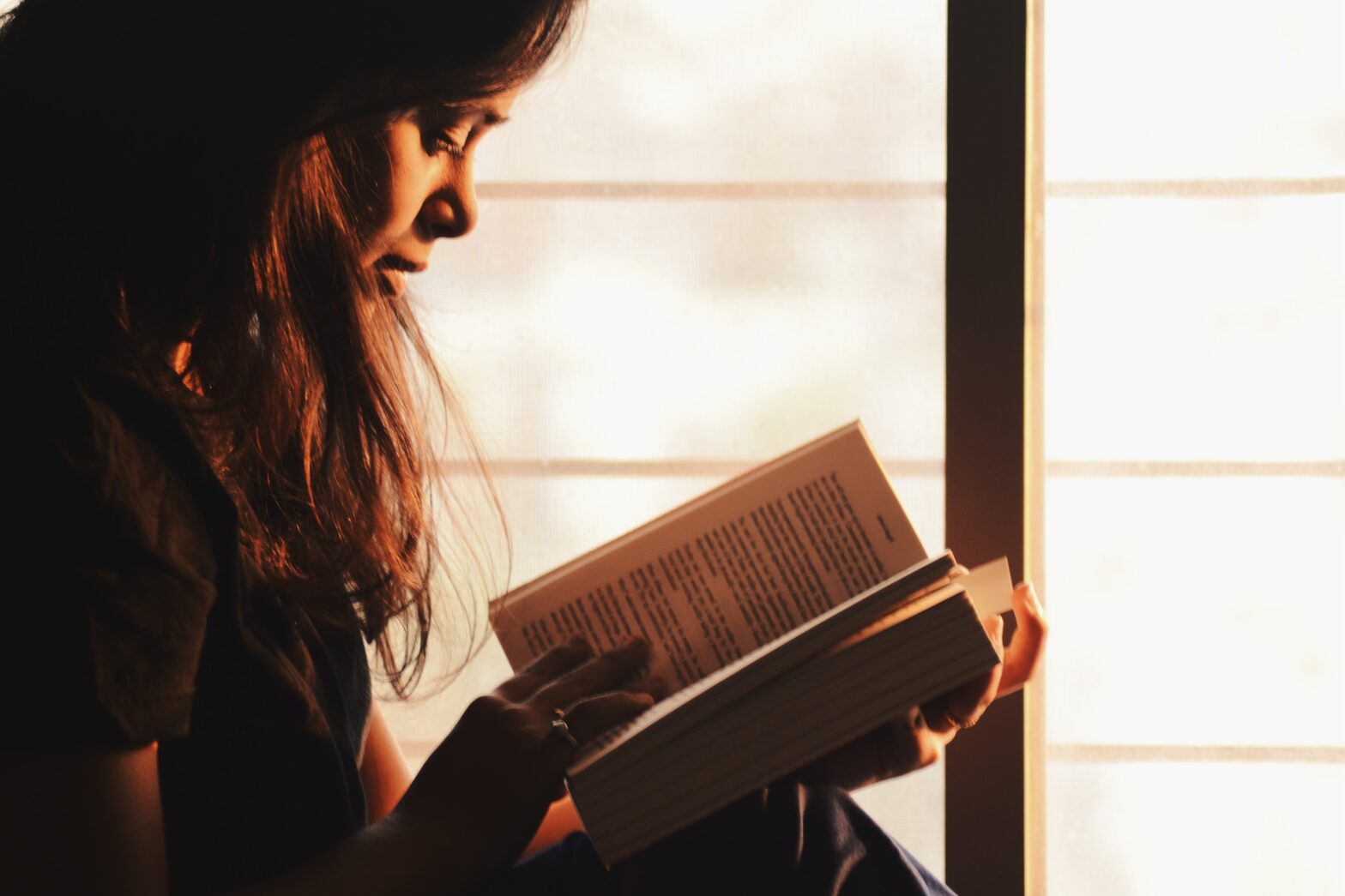 A girl reading a book beside the window.